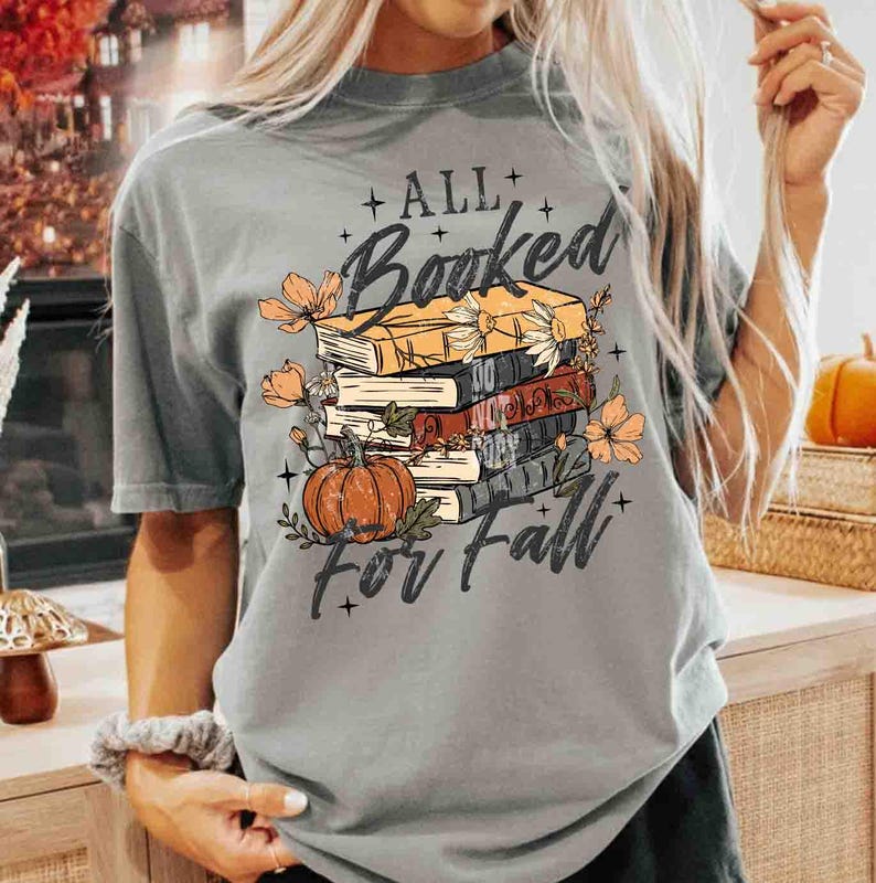 May include: Gray t-shirt with the text "ALL Booked For Fall" in a script font. The graphic features a stack of books, a pumpkin, and floral accents in orange and brown.