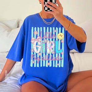 May include: A blue t-shirt featuring a graphic print with vertical teal stripes and the text "Little Girl Big attitude". The design includes a patterned bow, small flowers, and a yellow smiley face icon in a colourful, multi-toned aesthetic.