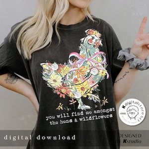 May include: A black t-shirt with a floral design of a chicken wearing a pink and blue ribbon. The text "you will find me amongst the hens & wildflowers" is printed below the chicken. The text "digital download" is printed at the bottom of the shirt. The text "DESIGNED BY Kstudio" is printed at the bottom right corner of the shirt.