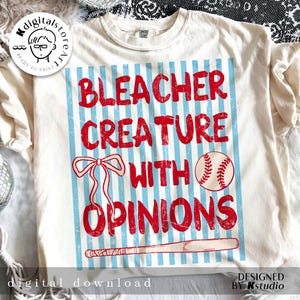 May include: Cream-colored t-shirt with a distressed graphic. The graphic features the text "BLEACHER CREATURE WITH OPINIONS" in red, set against a blue and white striped background, with baseball-themed illustrations.