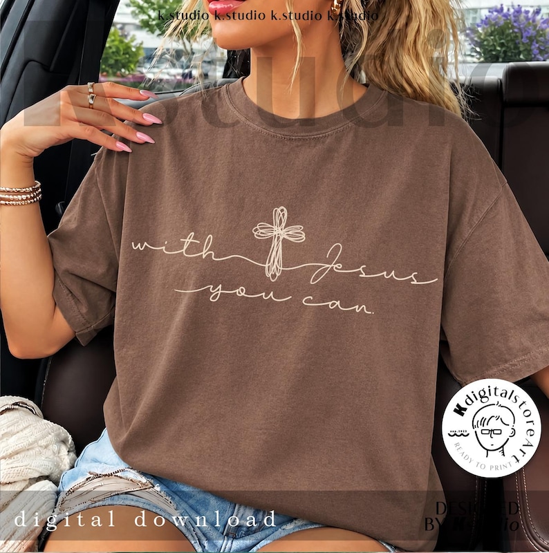 Op de afbeelding: Een bruin T-shirt met korte mouwen voorzien van een witte lijntekening van een kruis en de cursieve tekst "with Jesus you can". Het ontwerp is gecentreerd op de borst van het kledingstuk, dat wordt gedragen met een blauwe spijkerbroek.
