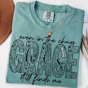 May include: A seafoam green t-shirt with the words "even in the chaos, Grace still finds me" in black script. The word "GRACE" is filled with a floral pattern. A silver necklace with a small pendant rests on the shirt.
