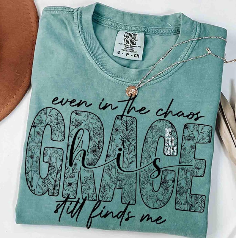 May include: A seafoam green t-shirt with the words "even in the chaos, Grace still finds me" in black script. The word "GRACE" is filled with a floral pattern. A silver necklace with a small pendant rests on the shirt.
