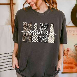 May include: Dark gray t-shirt with the word "mama" in white script. The letters of "mama" are filled with various patterns, including leopard print, black and white spots, and checkered designs. The shirt has a relaxed fit.