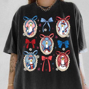 Op de afbeelding: Zwart T-shirt met een patriottisch ontwerp. Het heeft cartoon ganzen in ovale frames, elk gekleed in rood, wit en blauw, met voorwerpen als een fakkel en een zonnebril. Het shirt heeft een ronde hals en korte mouwen.