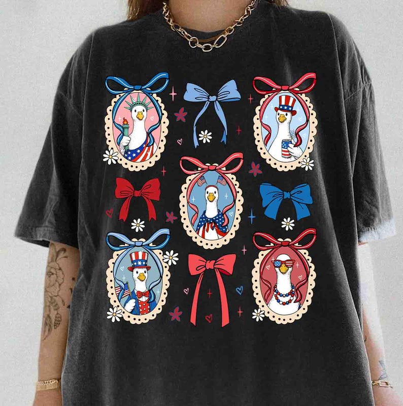 Op de afbeelding: Zwart T-shirt met een patriottisch ontwerp. Het heeft cartoon ganzen in ovale frames, elk gekleed in rood, wit en blauw, met voorwerpen als een fakkel en een zonnebril. Het shirt heeft een ronde hals en korte mouwen.
