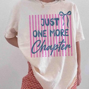 Puede incluir: Camiseta color crema con el texto "JUST ONE MORE Chapter" en escritura azul. El texto est&aacute; sobre un rect&aacute;ngulo a rayas rosas y blancas, con un lazo azul a la derecha. La camiseta es de estilo casual.