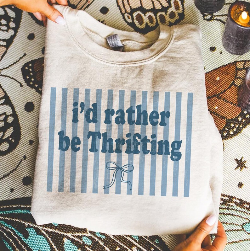 May include: A cream-colored sweatshirt with the text "I'd rather be Thrifting" in blue, set against a backdrop of vertical blue stripes. A blue bow graphic is below the text. The sweatshirt is folded.