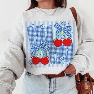 May include: A light gray sweatshirt with a blue gingham "MOM" design. The letters are decorated with red cherries, blue bows, and small white flowers. The background has blue vertical stripes.