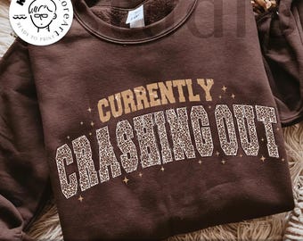 May include: A brown sweatshirt with the text "CURRENTLY CRASHING OUT" in a combination of tan and leopard print. The words are arched across the front of the garment. The phrase "digital download" is at the bottom.