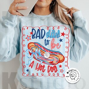 May include: Light blue sweatshirt with a graphic that says "BAD day to be A HOT DOG". The graphic features a colorful hot dog with a blue bow, red stars, and blue and red text.
