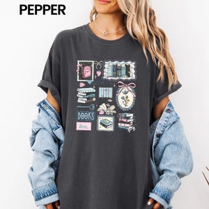 May include: Dark gray t-shirt with a colorful graphic of books, stamps, and floral elements. The word "BOOKS" is printed in a blue font, and the word "love" is printed in a pink font. The shirt has short sleeves.