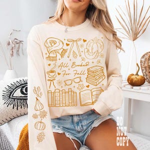 May include: Cream-colored long-sleeve shirt with a fall-themed graphic. The design includes a scarf, bow, pumpkin, books, coffee cup, glasses, and the text "All Booked For Fall".