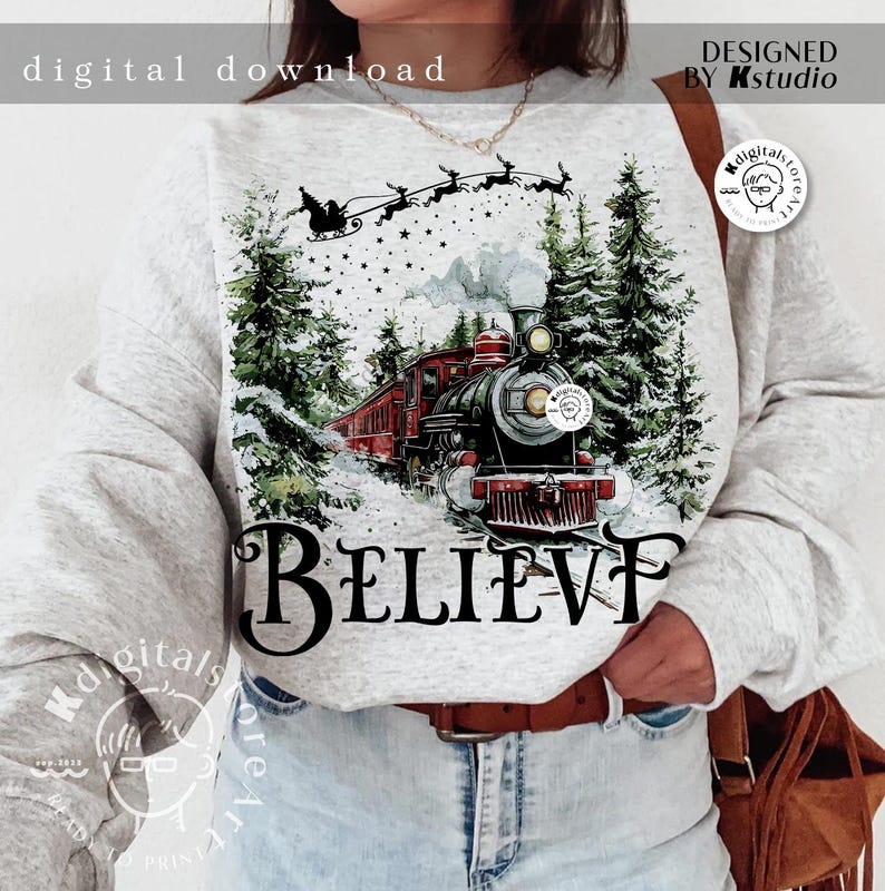 May include: A gray sweatshirt with a watercolor design of a red train traveling through a snowy forest. The train is pulling a string of boxcars. The words "BELIEVE" are printed below the train. Reindeer are flying in the sky above the train.