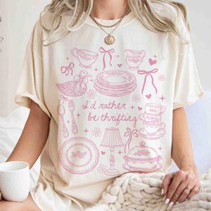 May include: Cream-colored t-shirt with a pink graphic of vintage dishware, a duck, and the text "I'd rather be thrifting." The design includes teacups, plates, and a lamp. The shirt has short sleeves and a relaxed fit.
