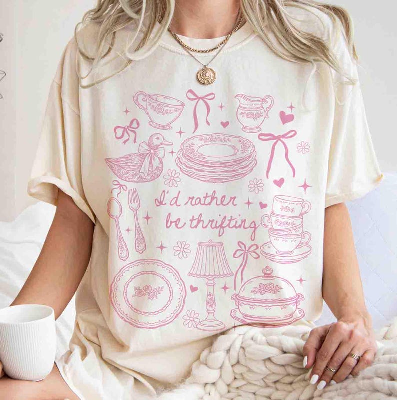 May include: Cream-colored t-shirt with a pink graphic of vintage dishware, a duck, and the text "I'd rather be thrifting." The design includes teacups, plates, and a lamp. The shirt has short sleeves and a relaxed fit.