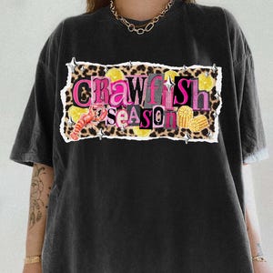 May include: Black t-shirt with a graphic that reads "Crawfish Season." The design includes a crawfish, lemon slices, and corn on the cob, set against a leopard print background. The text is in pink and yellow.