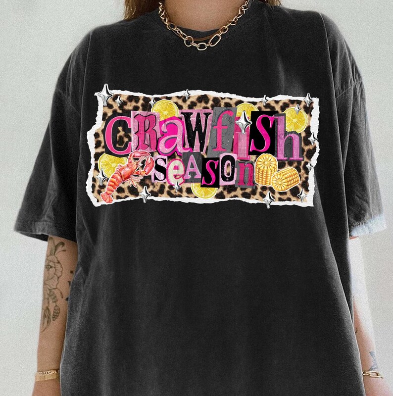 May include: Black t-shirt with a graphic that reads "Crawfish Season." The design includes a crawfish, lemon slices, and corn on the cob, set against a leopard print background. The text is in pink and yellow.
