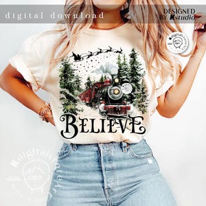 May include: A white t-shirt with a Christmas-themed design featuring a red and black train traveling through a snowy forest. The train is pulling a string of Christmas lights and the text "BELIEVE" is printed below the train.