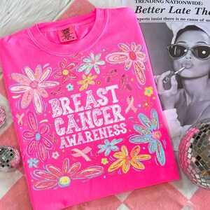 May include: A bright pink t-shirt with the words "BREAST CANCER AWARENESS" in white, surrounded by colorful floral and ribbon graphics. The shirt is made of a soft material and has a crew neck.