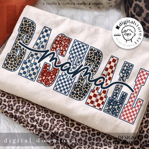 Op de afbeelding: Crèmekleurig T-shirt met de tekst "Baseball Mama" in een mix van patronen en kleuren. Het woord "Baseball" is in luipaardprint, geruit en gestippeld. Het woord "Mama" is in een sierlijk lettertype.