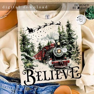 May include: A beige sweatshirt with a watercolor illustration of a train traveling through a snowy forest. The train is red and black with smoke coming from the chimney. The text "BELIEVE" is printed in black below the train. Santa Claus is flying in the sky above the train with reindeer.