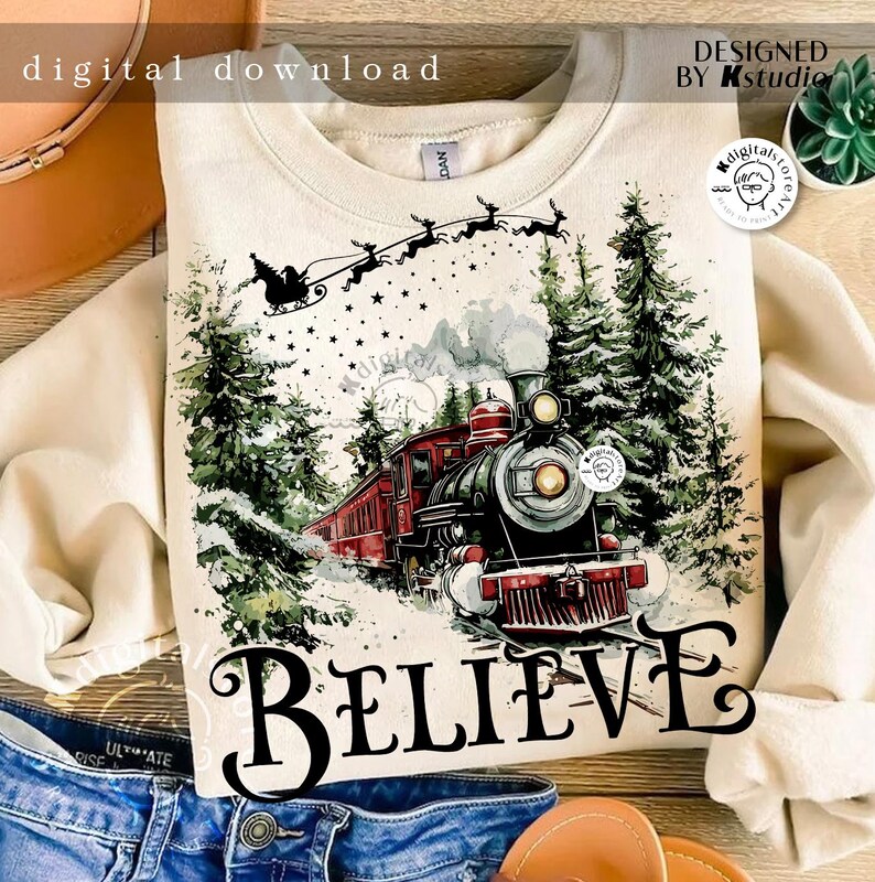 May include: A beige sweatshirt with a watercolor illustration of a train traveling through a snowy forest. The train is red and black with smoke coming from the chimney. The text "BELIEVE" is printed in black below the train. Santa Claus is flying in the sky above the train with reindeer.
