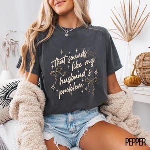 May include: Dark gray t-shirt with the phrase "that sounds like my husband's problem" in white script and leopard print accents. The shirt has a relaxed fit and is worn with denim shorts.
