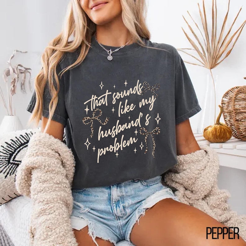 May include: Dark gray t-shirt with the phrase "that sounds like my husband's problem" in white script and leopard print accents. The shirt has a relaxed fit and is worn with denim shorts.