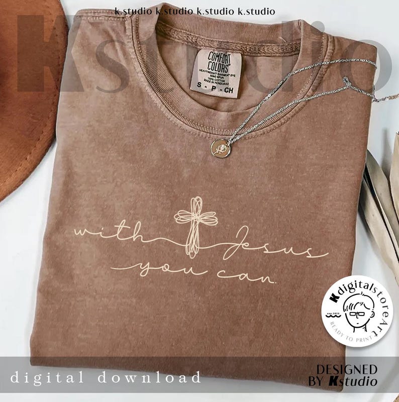 Op de afbeelding: Een bruin T-shirt met korte mouwen, voorzien van een wit lijntekening-kruis en de tekst "with Jesus you can". Een zilverkleurige ketting met een kleine ronde hanger rust op de halslijn. Het shirt heeft een Comfort Colors-label in de kraag.