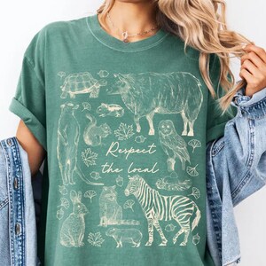 May include: Green t-shirt with a cream-colored illustration of various animals, including a zebra, owl, and squirrel. The text "Respect the local" is centered below the animals. The shirt has short sleeves and a relaxed fit.
