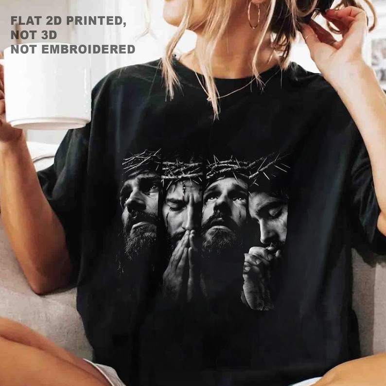 May include: Black t-shirt featuring a grayscale graphic of four faces, each wearing a crown of thorns. The text "FLAT 2D PRINTED, NOT 3D, NOT EMBROIDERED" is printed at the top. The shirt is a casual style.