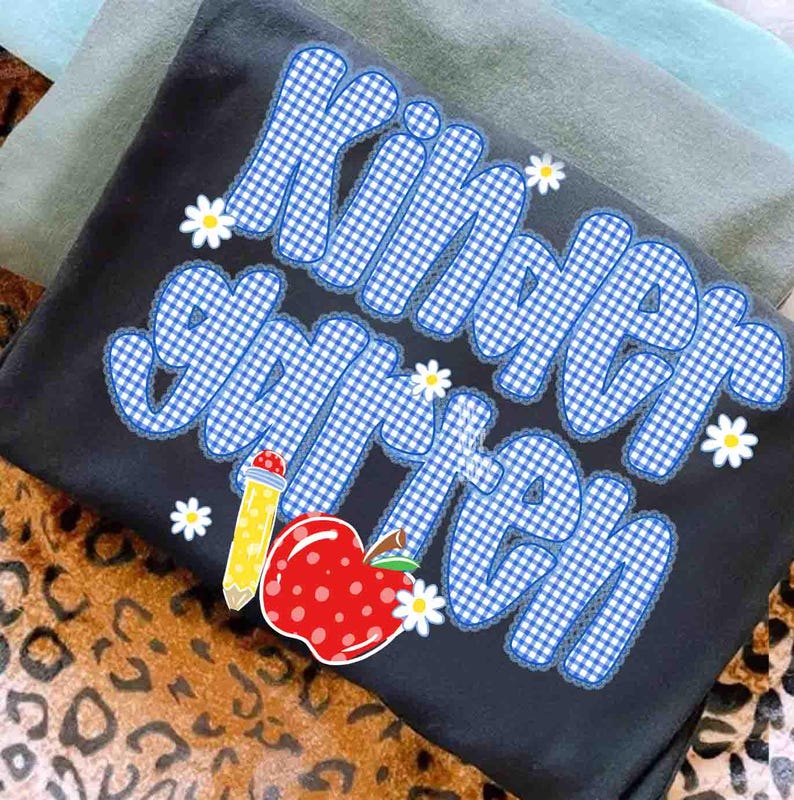 以下が含まれることがあります： 青と白のギンガムチェック柄でKindergartenという文字がプリントされた黒いTシャツ。デザインには、黄色い鉛筆、白い水玉模様の赤いリンゴ、そして文字の周りに散らばった小さな白いデイジーの花が含まれています。
