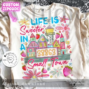 May include: A cream-coloured t-shirt featuring a vibrant graphic. The design says "Life is Sweeter in a Small Town" with houses, flowers, strawberries, and the number "38863."