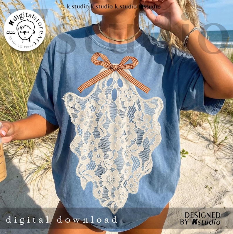 May include: A blue short-sleeved t-shirt featuring a large graphic of a ghost shape composed of cream-colored floral lace. The ghost graphic is topped with a brown and white gingham patterned bow. The shirt has a crew neckline and a relaxed fit.