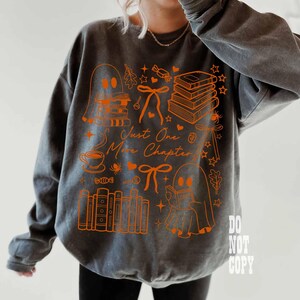 May include: Gray sweatshirt with an orange graphic design featuring ghosts, books, bows, and the text "Just One More Chapter." The design includes a coffee cup, candy, and stars. The sweatshirt is a comfortable, casual style.