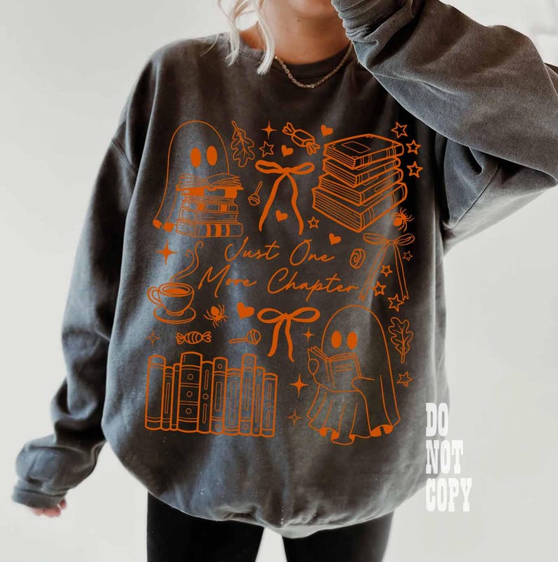 May include: Gray sweatshirt with an orange graphic design featuring ghosts, books, bows, and the text "Just One More Chapter." The design includes a coffee cup, candy, and stars. The sweatshirt is a comfortable, casual style.