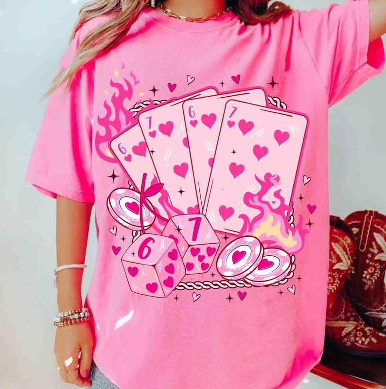 May include: Pink t-shirt featuring a graphic of playing cards, dice, and poker chips with heart symbols. The design includes flames and decorative elements. The shirt is a solid pink color.
