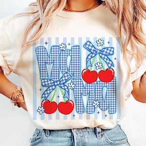 May include: Cream-colored t-shirt featuring the word "MOM" in a blue gingham pattern with red cherries and blue bows. The design includes white flowers and blue vertical stripes. A casual and cheerful design.