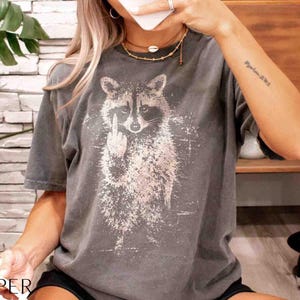 May include: Dark gray t-shirt featuring a distressed graphic of a raccoon giving the middle finger. The raccoon is in white, with a splash effect. The shirt has short sleeves and a crew neck.