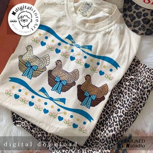 May include: Cream-colored t-shirt with a whimsical chicken design. Three chickens are in baskets with blue gingham bows, accented by blue ribbons, hearts, and floral details. The words "digital download" are visible.