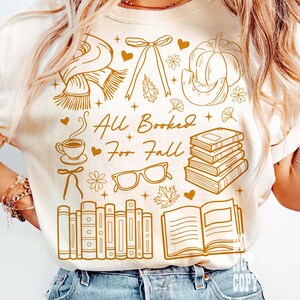 May include: Cream-colored t-shirt with a brown graphic design featuring fall-themed illustrations. The design includes a scarf, bow, pumpkin, coffee cup, glasses, books, and the text "All Booked For Fall."