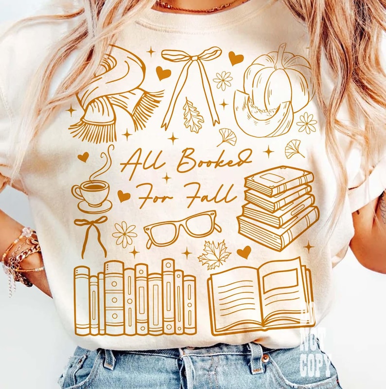 May include: Cream-colored t-shirt with a brown graphic design featuring fall-themed illustrations. The design includes a scarf, bow, pumpkin, coffee cup, glasses, books, and the text "All Booked For Fall."