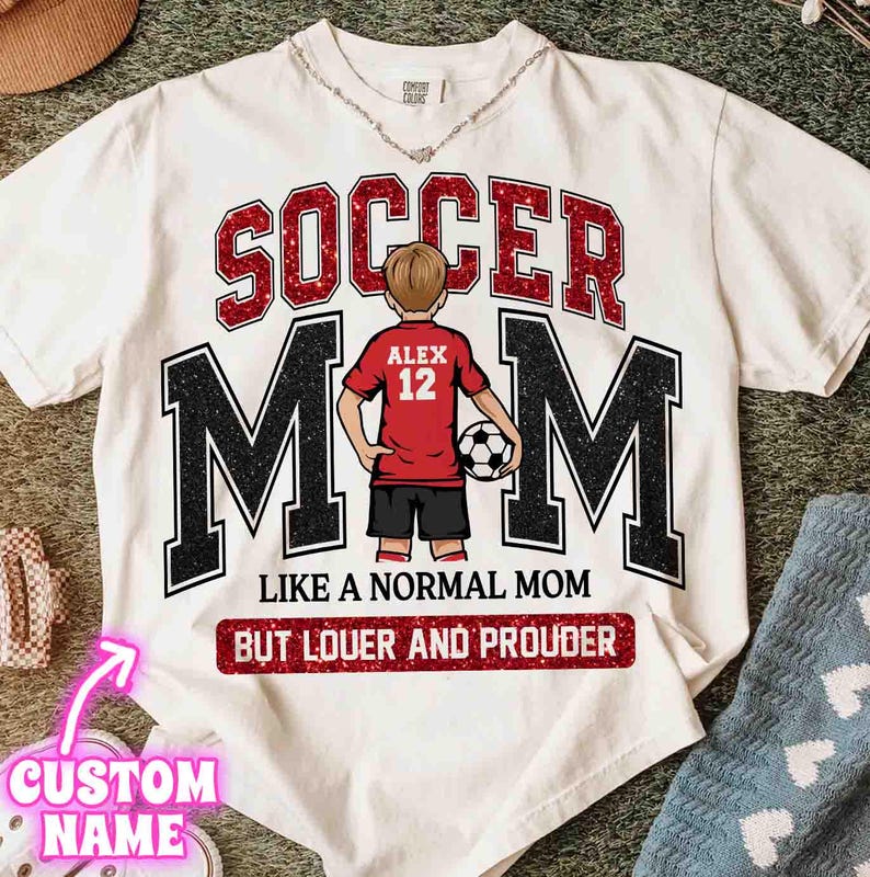 May include: White t-shirt with a soccer theme. The shirt features the words "SOCCER MOM" in red and black, with an illustration of a soccer player. Text below reads "LIKE A NORMAL MOM BUT LOUDER AND PROUDER."