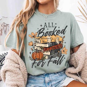 May include: A light green t-shirt with the text "All Booked For Fall" featuring a stack of books, a pumpkin, and floral accents. The design is in black, orange, and yellow.