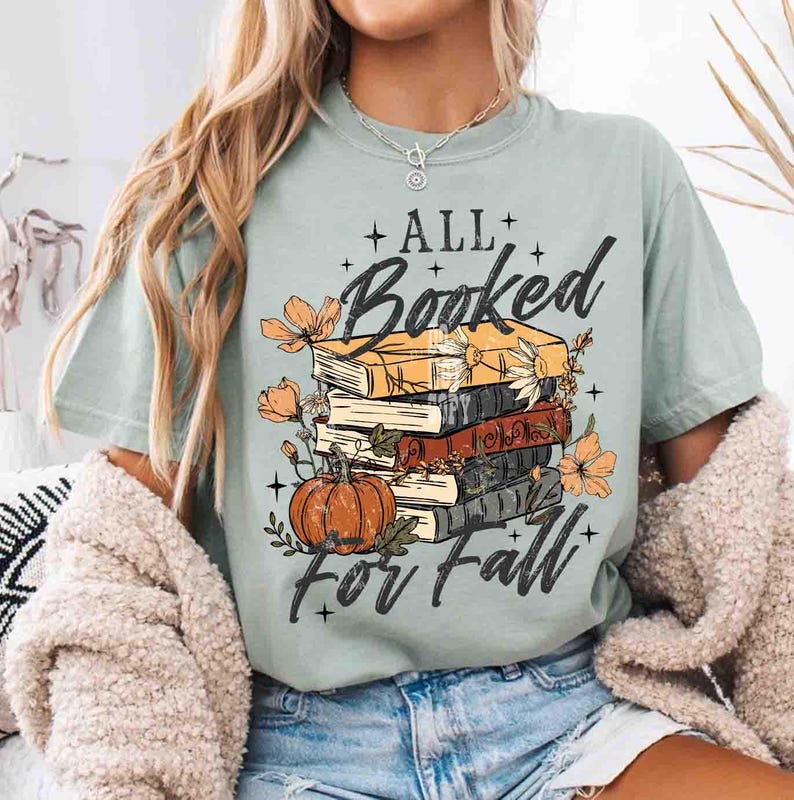 May include: A light green t-shirt with the text "All Booked For Fall" featuring a stack of books, a pumpkin, and floral accents. The design is in black, orange, and yellow.