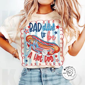 May include: Cream-colored t-shirt with a graphic that reads "BAD day to be A Hot Dog." The design features a hot dog illustration with red, blue, and pink accents, set against a striped background with stars.