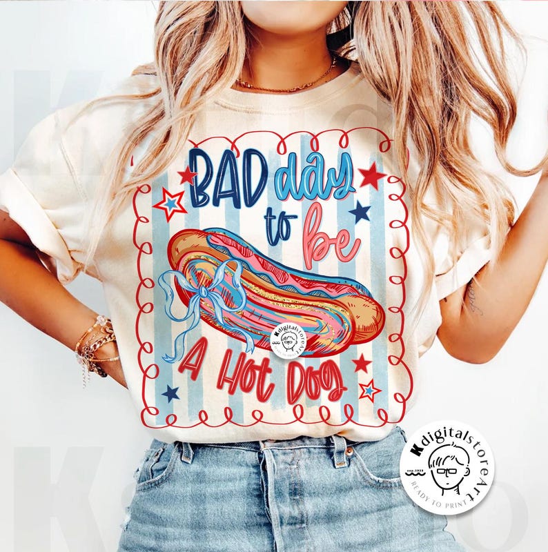 May include: Cream-colored t-shirt with a graphic that reads "BAD day to be A Hot Dog." The design features a hot dog illustration with red, blue, and pink accents, set against a striped background with stars.