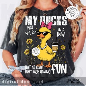 Puede incluir: Camiseta gris oscuro con un gr&aacute;fico de un pato amarillo con gafas de sol, un lazo rosa y un collar de perlas, sosteniendo una taza de caf&eacute;. El texto dice "MY DUCKS MAY NOT BE IN A ROW BUT AT LEAST THEY ARE HAVING FUN."