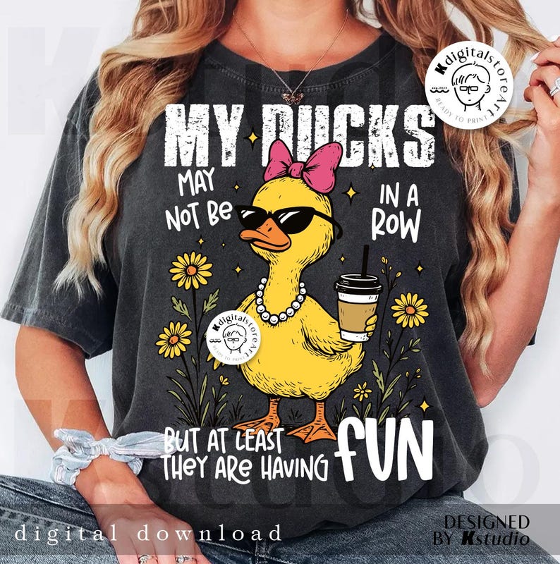 Puede incluir: Camiseta gris oscuro con un gr&aacute;fico de un pato amarillo con gafas de sol, un lazo rosa y un collar de perlas, sosteniendo una taza de caf&eacute;. El texto dice "MY DUCKS MAY NOT BE IN A ROW BUT AT LEAST THEY ARE HAVING FUN."